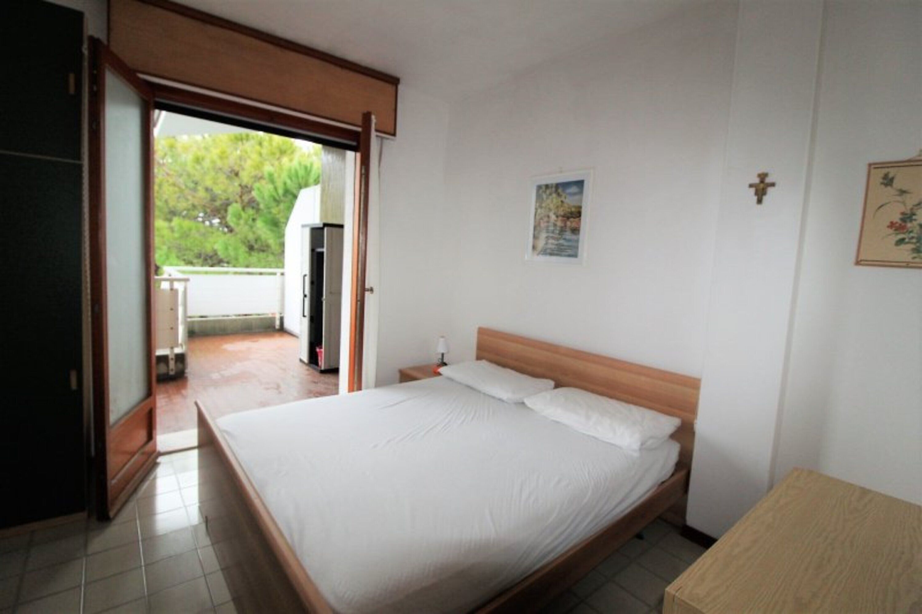 Un dormitorio con cama, una mesita de noche, una pequeña mesa, una puerta que da a un balcón y una cruz colgada en la pared.