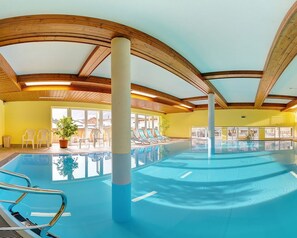 Una piscina interior con vigas de madera, paredes de vidrio transparente y sillas de descanso.