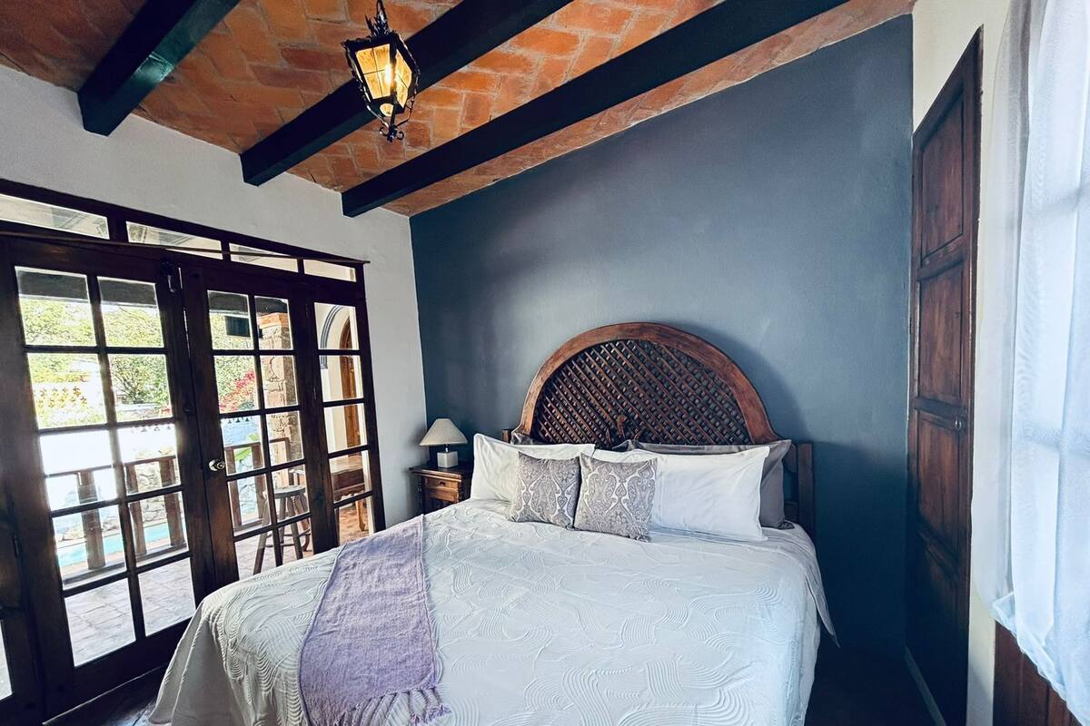 A bedroom with a large bed, a wooden headboard, a nightstand, and a window with a view of greenery.