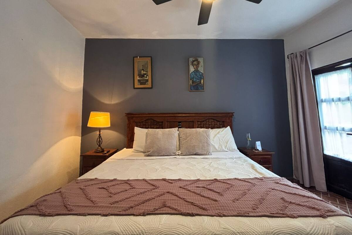 A bedroom with a bed, a ceiling fan, a nightstand with a lamp, two framed pictures on the wall, and a window with curtains.