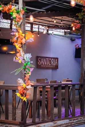 Et restaurantinteriør med et skilt der viser 'Santoria Restaurante', træborde, stole og dekorative blomsterarrangementer.