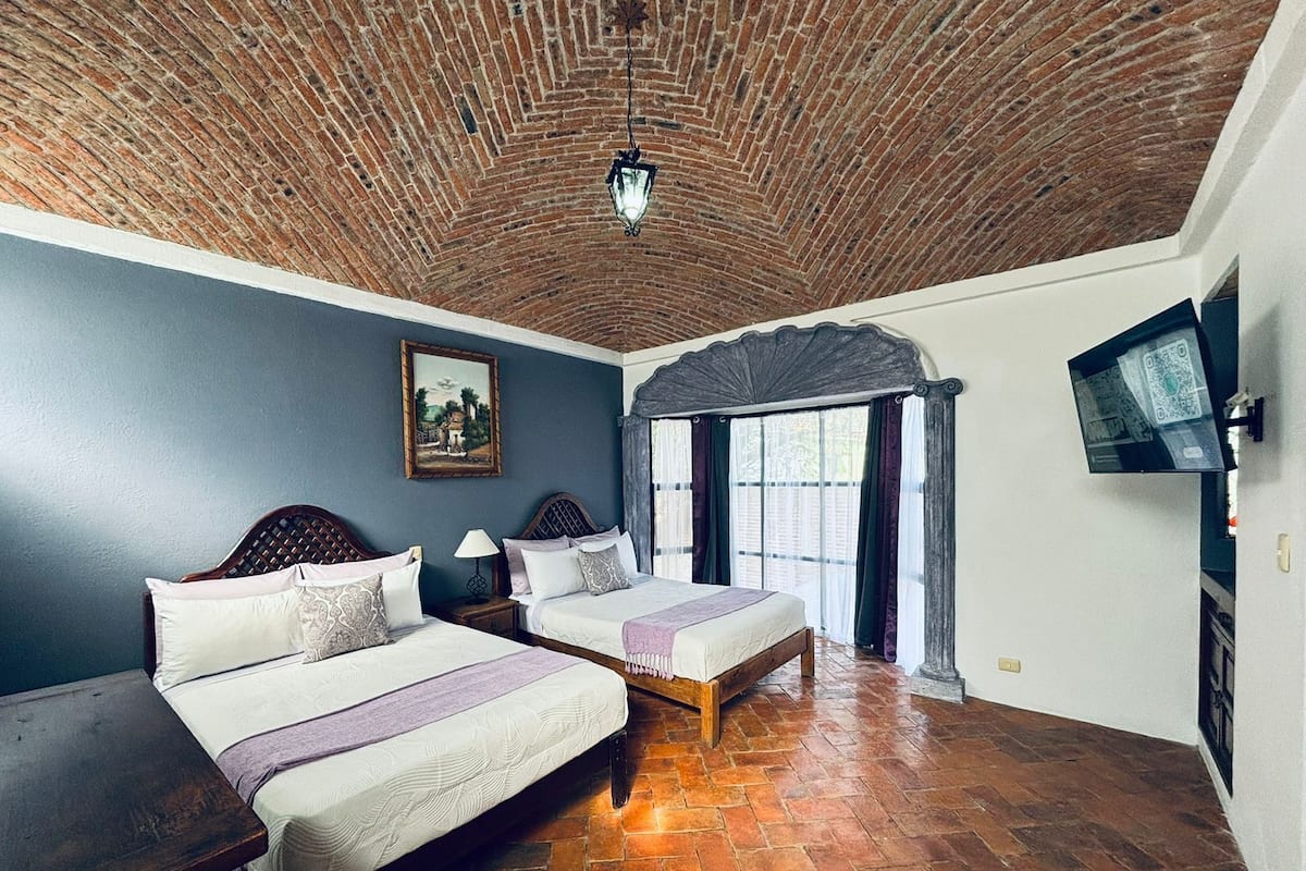 A room with two beds, a brick ceiling, a painting on the wall, and a TV mounted on the wall.