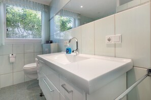A modern bathroom with a large white sink, a toilet, and a window with blinds.