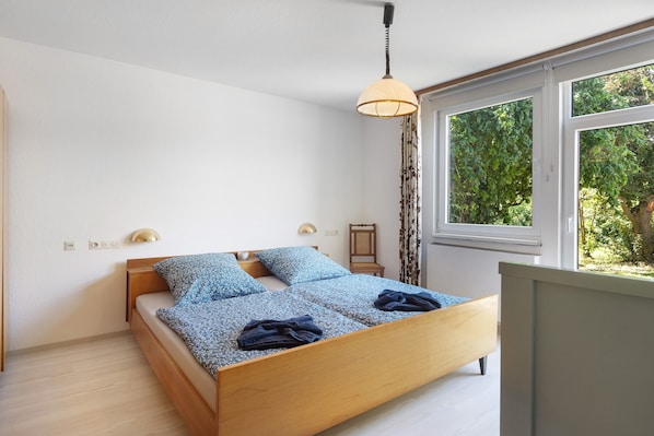 Ein Schlafzimmer mit einem hölzernen Bett, einer Pendelleuchte, einem Nachttisch und einem Fenster mit Blick auf Grünflächen.