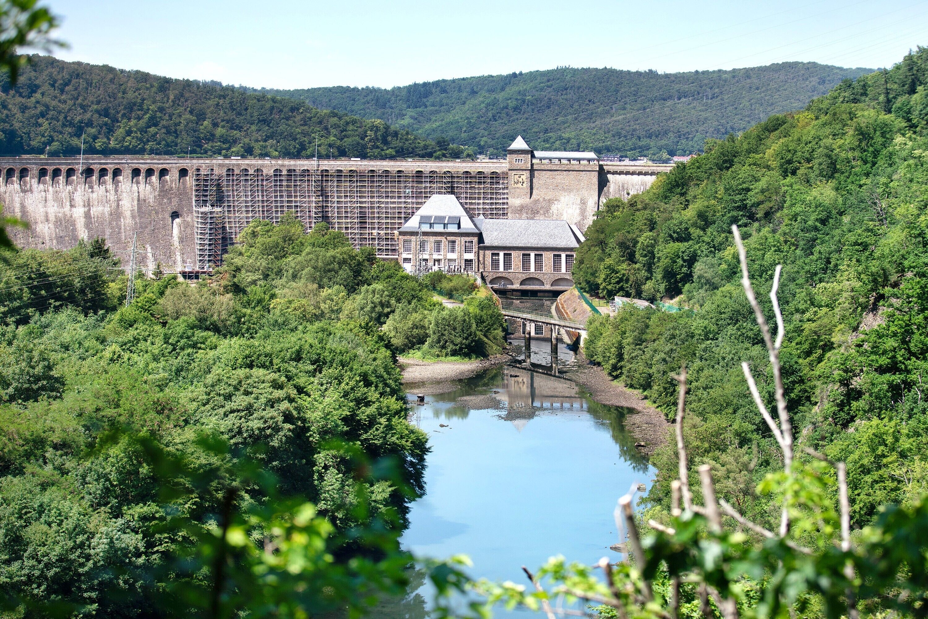 Un'imponente diga con un ponte, un edificio con una torre e un fiume circondato da vegetazione.