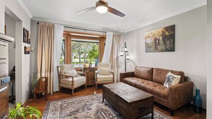 Un salon comprenant un canapé, des fauteuils, une table basse, un ventilateur de plafond, une fenêtre avec des rideaux et un tableau accroché au mur.