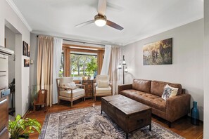 A living room with a sofa, chairs, a coffee table, a ceiling fan, a window with curtains, and a painting on the wall.