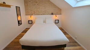 Ein Schlafzimmer mit schrägen Wänden, einem Bett mit weißer Bettwäsche, zwei an der Wand befestigten Nachttischen mit Lampen und einem Holzboden.