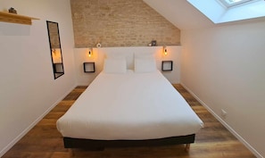 Une chambre avec un plafond en pente, un lit avec une literie blanche, deux tables de chevet fixées au mur, chacune avec une lampe, et un plancher en bois.