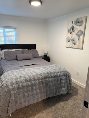 A bedroom with a bed, a nightstand, a lamp, a painting, and a window with blinds. - Buckeye Lake Retreat w/ Fire pit/ Sleeps 8 (Thornville)
