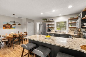 A modern kitchen with a large island, stainless steel appliances, and a dining area with wooden furniture. - Big Sky Escape | Ski In/Ski Out + Mountain Views (Big Sky)