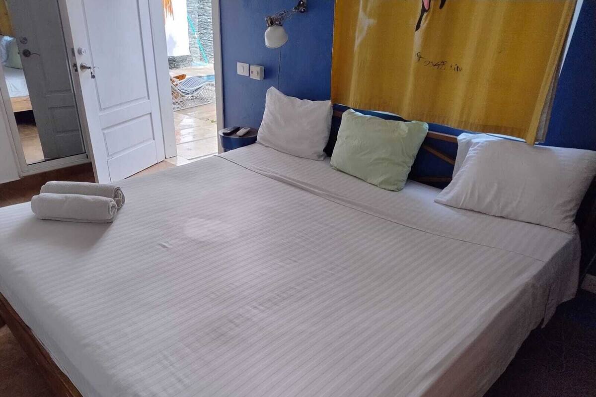 Un lit bien fait, avec du linge de lit blanc, un mur bleu et un rideau jaune.