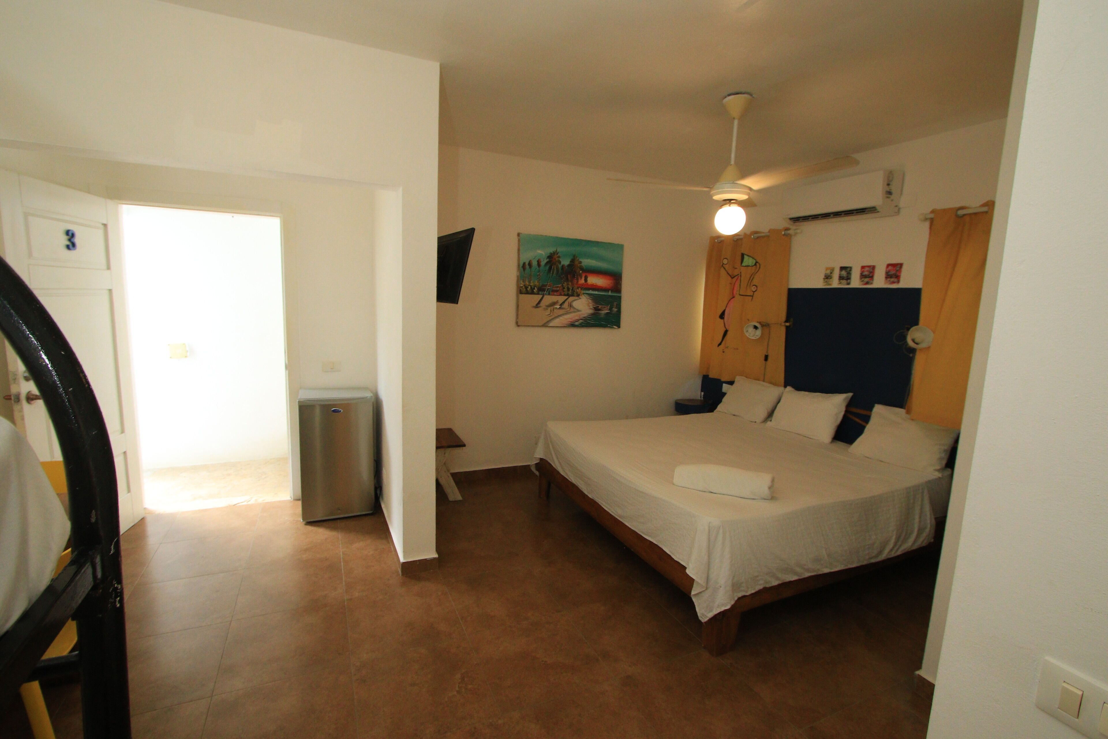 Une chambre à coucher avec un lit, un ventilateur de plafond, une petite poubelle et une porte donnant sur une autre pièce.