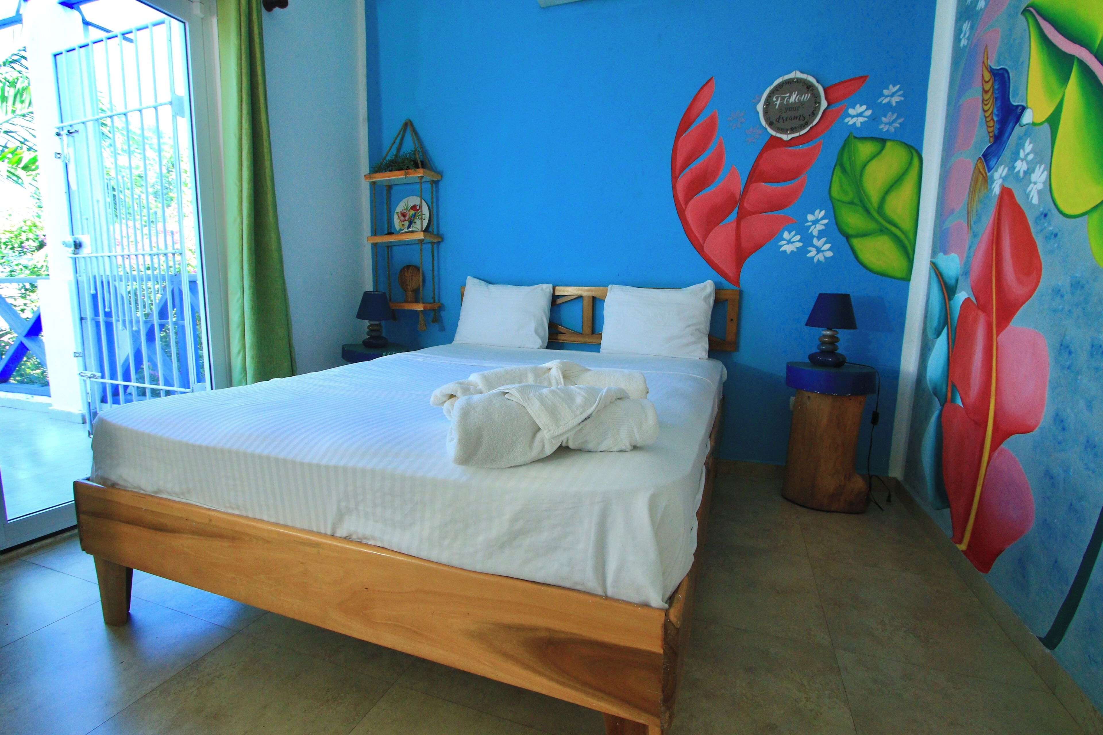 A bedroom with a wooden bed, white bedding, a small shelf with decorative items, and a colorful wall mural.