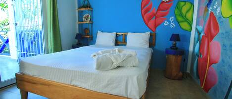 A bedroom with a wooden bed, white bedding, a small shelf with decorative items, and a colorful wall mural.