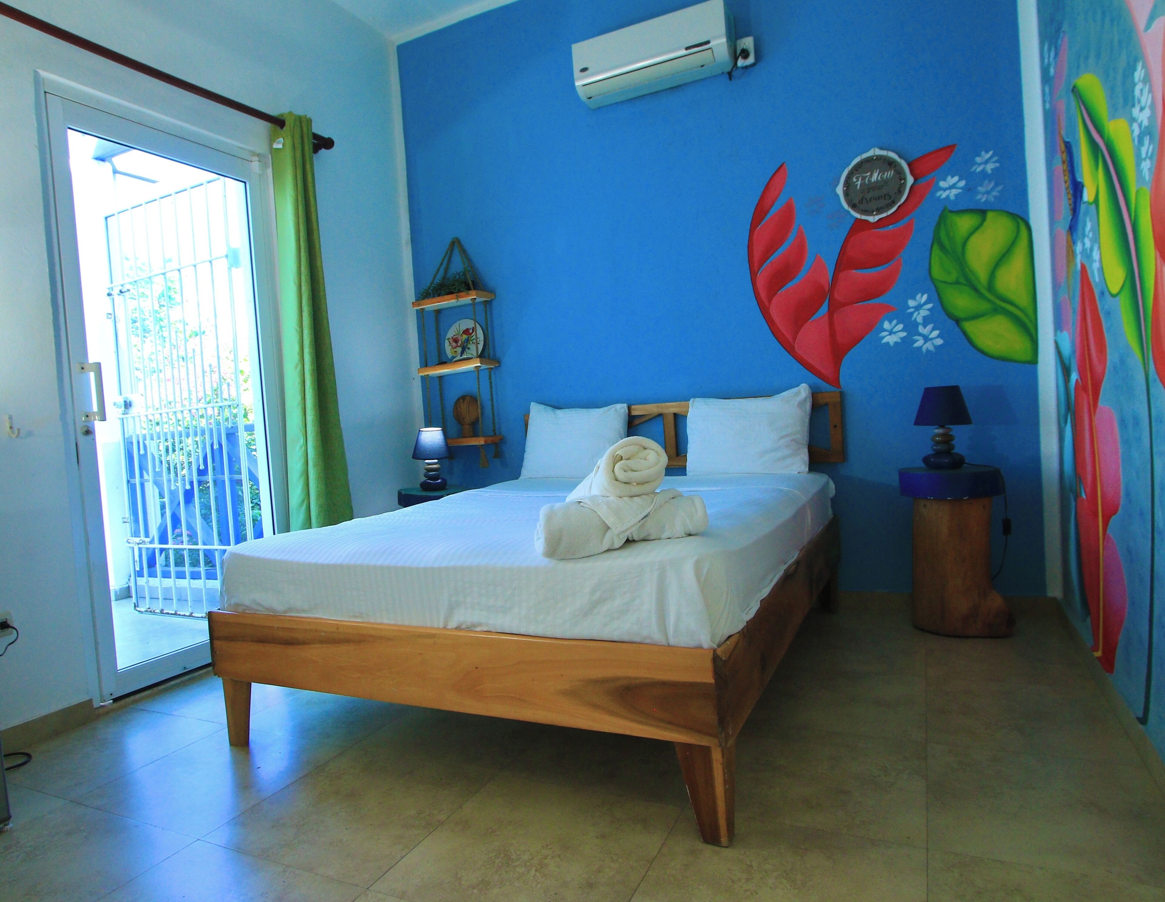 A room with a bed, a wall-mounted air conditioner, a small shelf with decorative items, and a colorful mural.