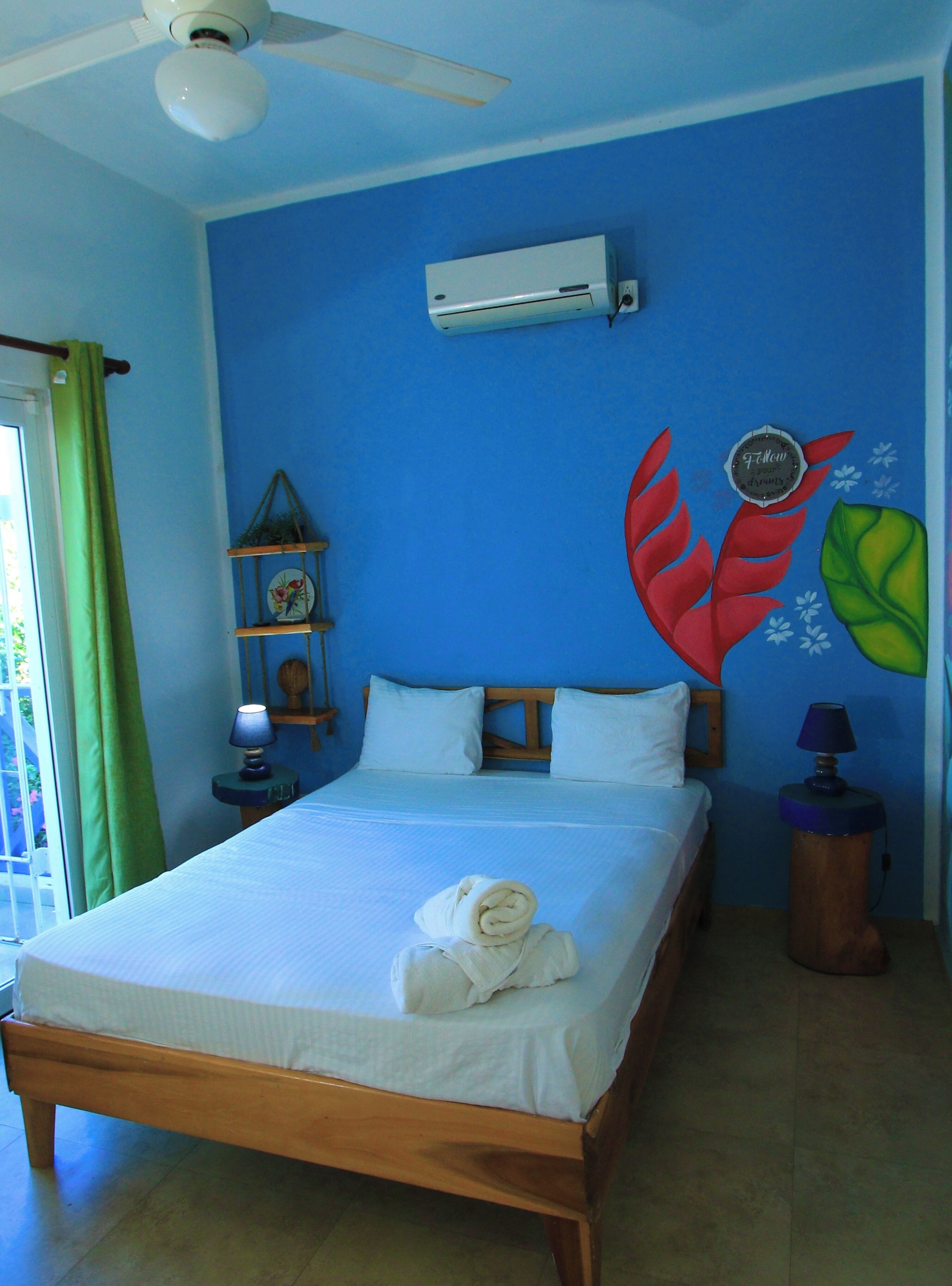 A bedroom with a bed, a ceiling fan, an air conditioner, a nightstand with a lamp, and a wall decoration.