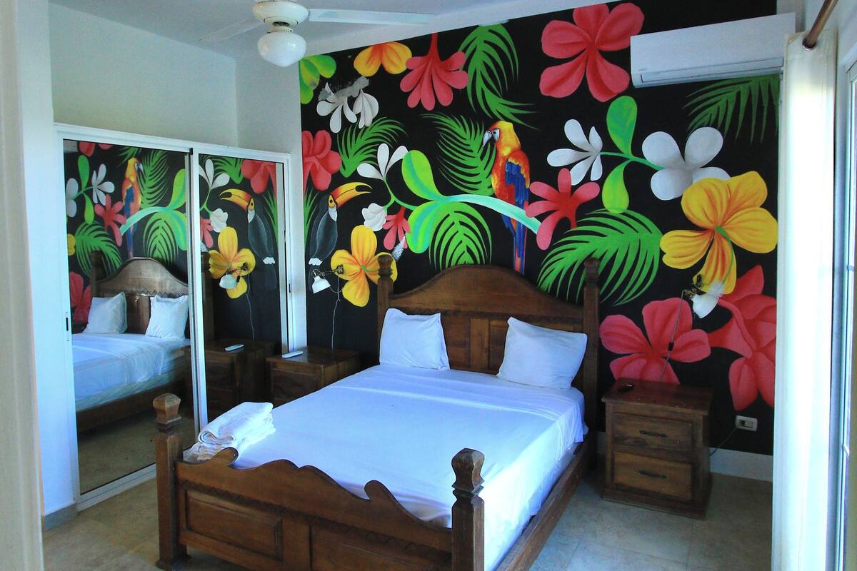 Une chambre à coucher comprenant un lit, une tête de lit en bois, des tables de chevet, un ventilateur de plafond et un mur recouvert d’un papier peint à motifs floraux tropicaux.