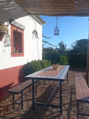 Eine Terrasse mit Tisch und Bänken, einem hängenden Vogelkäfig und einem Gebäude mit rotem Fenster und dem Schild 'ANTONIO'.