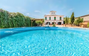 Ein Pool mit glasklar blauem Wasser, ein Gebäude mit weißer Fassade und roten Akzenten im Hintergrund sowie ein grüner Hügel.
