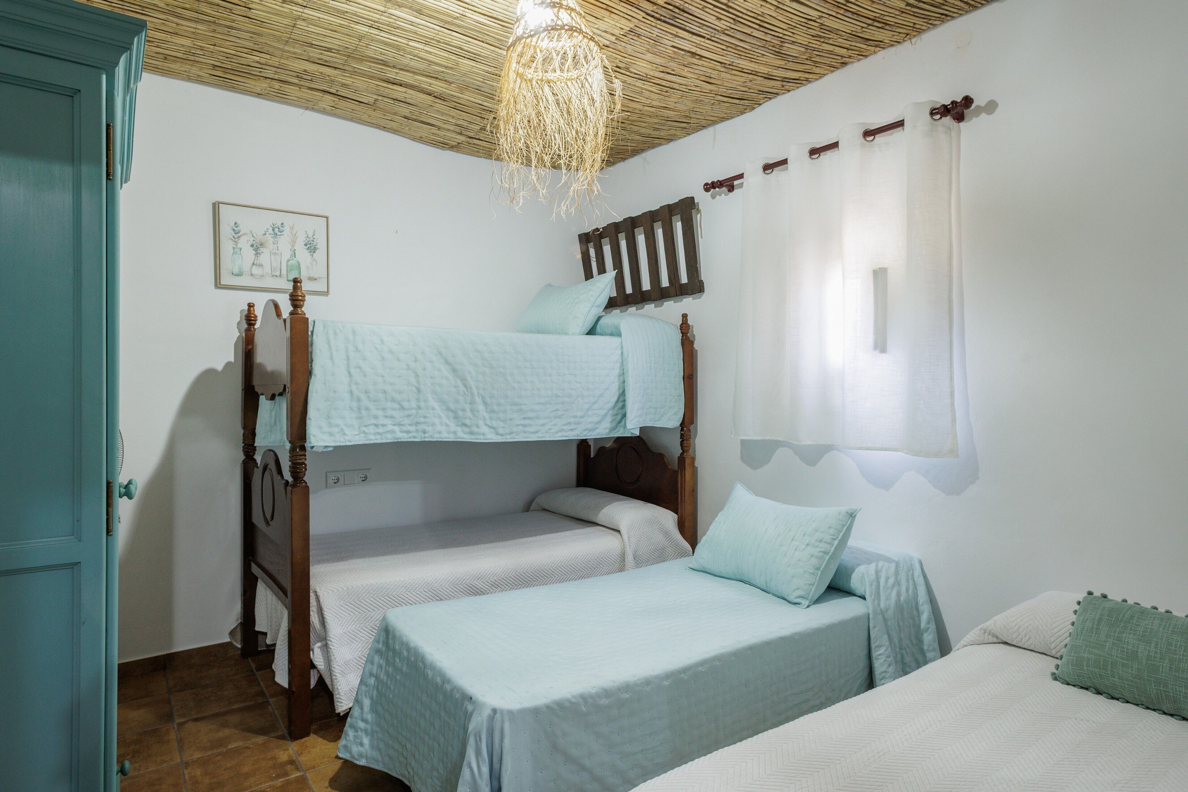 Une pièce avec un lit superposé, un lit simple, une porte en bois, un luminaire suspendu au plafond fait de fibres tressées et une étagère fixée au mur.