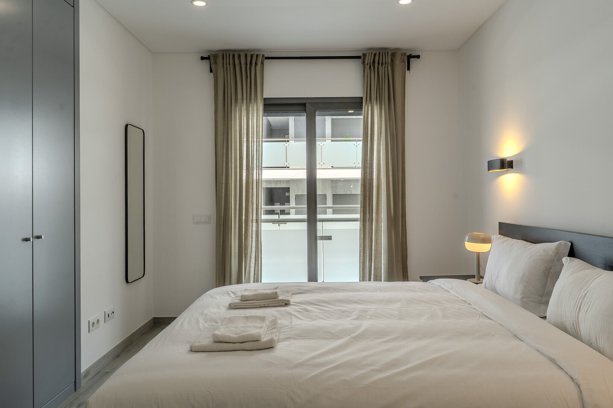 Ein modernes Hotelzimmer mit einem großen Bett, Blick auf ein Gebäude durch eine Glastür, einer Nachttischlampe und einem Kleiderschrank.