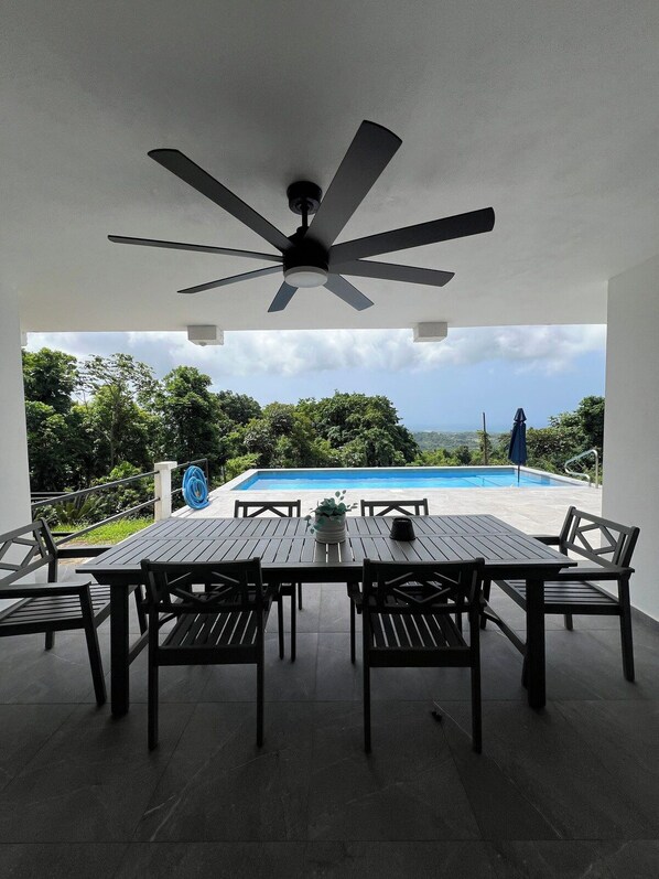 Une terrasse extérieure avec une table et des chaises, un ventilateur de plafond et une vue sur une piscine.
