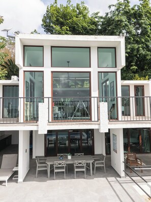Un bâtiment moderne à deux étages, doté de grandes fenêtres, d’un balcon avec une rambarde en métal et d’un patio aménagé avec des tables et des chaises.