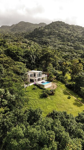 Une maison moderne avec une piscine, entourée d’une végétation luxuriante et avec des collines en arrière-plan.