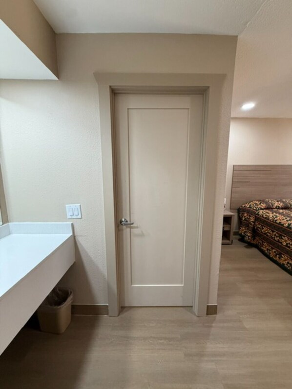 Une pièce avec une porte, une petite table de chevet, un lit recouvert d’une couvre-lit à motifs, une étagère blanche et une poubelle.