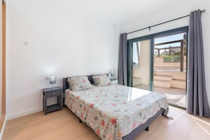 Une chambre à coucher avec un lit, des tables de chevet, une lampe et une vue sur l’extérieur, accessible par une porte vitrée.