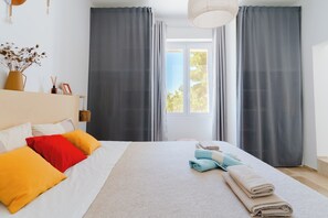 Ein ordentlich gemachtes Bett mit Kissen, ein Fenster mit Vorhängen und eine Vase mit Trockenblumen auf einem Nachttisch.