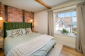 Ein Schlafzimmer mit Backsteinwand, einem grünen Kopfteil, einem großen Fenster mit Vorhängen und Blick auf ein Haus.