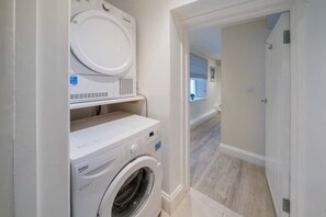 A laundry room with a front-loading washer and dryer, a small shelf, and a door leading to another room. - The Watch House Waterfront Home With Solent Views (Cowes)