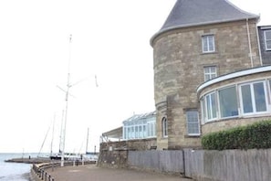 A stone building with a curved corner, a large window, and a wooden fence by the water. - The Watch House Waterfront Home With Solent Views (Cowes)