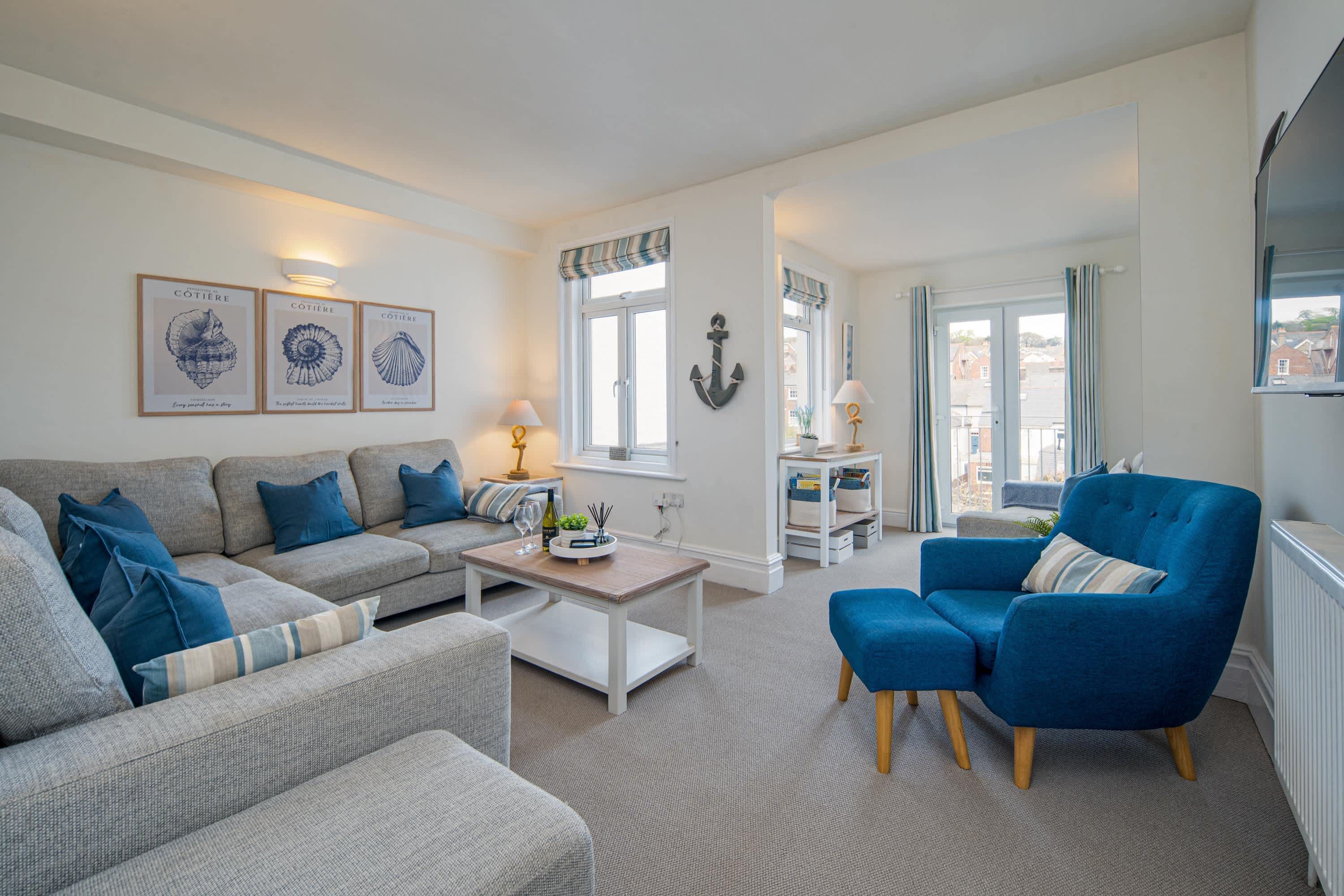 Ein Wohnzimmer mit Sofa, Sessel und Couchtisch. An der Wand hängen Bilderrahmen, ein Fenster mit Vorhängen und ein Blick ins Freie.