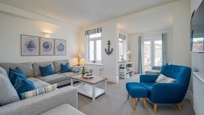 Una sala de estar con sofá, sillón y mesa de centro. Hay cuadros en la pared, una ventana con cortinas y vistas al exterior.