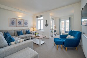 Ein Wohnzimmer mit Sofa, Sessel und Couchtisch. An der Wand hängen Bilderrahmen, ein Fenster mit Vorhängen und ein Blick ins Freie.