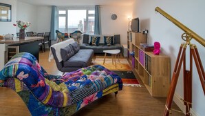 Ein Wohnzimmer mit einer Couch, einem bunten Sessel, einem kleinen Tisch, einem Bücherregal und einem Teleskop.