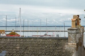Blick auf das Wasser mit einer Steinmauer, einem kleinen Gebäude mit gelbem Dach und mehreren Segelbooten im Hintergrund, die im Hafen liegen.