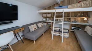 Ein Loft-Zimmer mit Fernseher, Schreibtisch, Sofa und einer Leiter zum Schlafplatz in der Erhöhung.