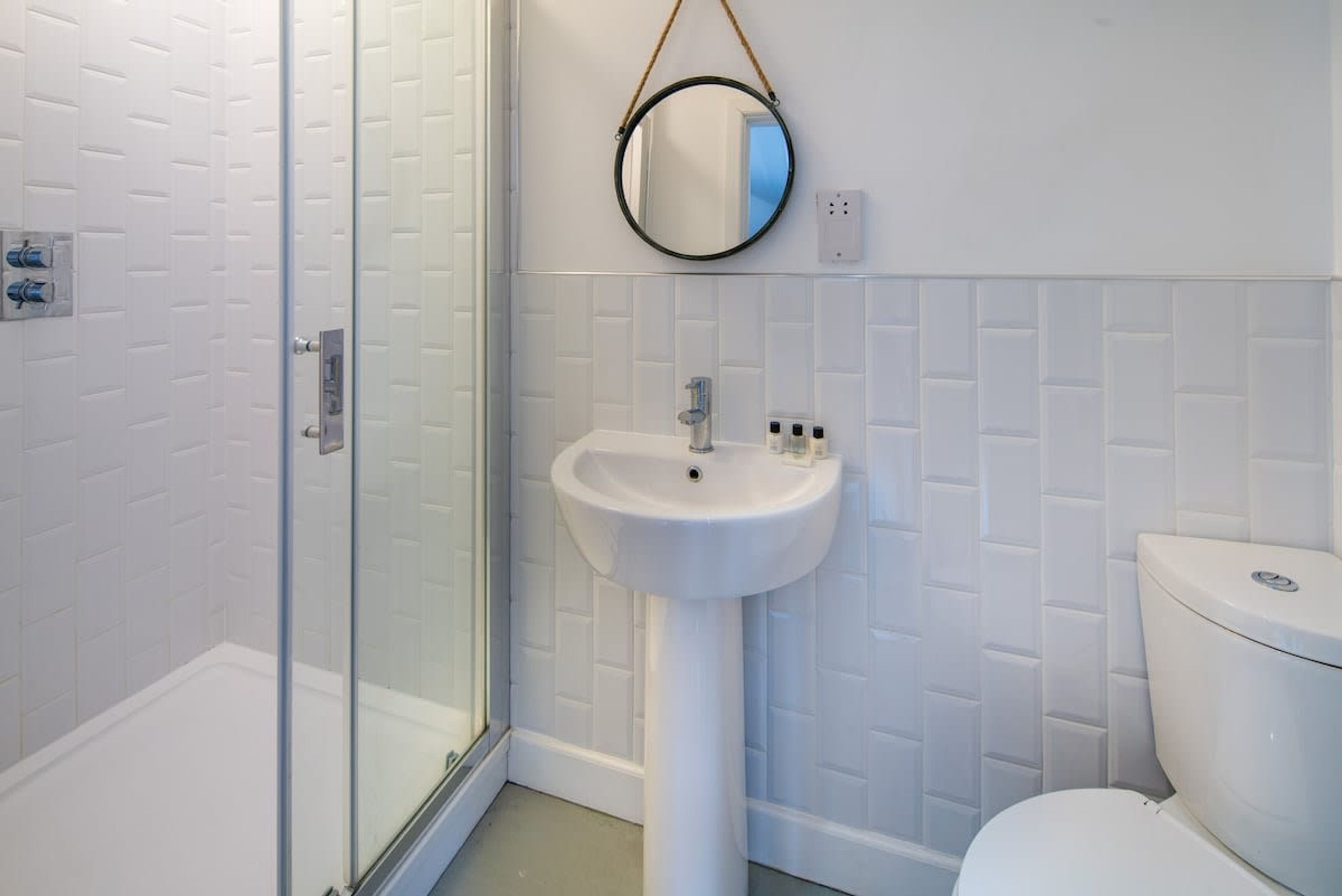 A bathroom with a glass shower enclosure, a pedestal sink, a toilet, and a round mirror on the wall.