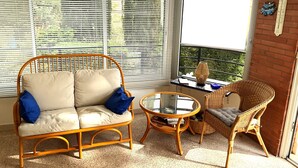 Une pièce comprenant un canapé en osier, un fauteuil en osier, une petite table ronde et une fenêtre avec des stores.