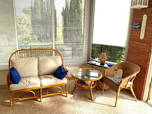 Ein Zimmer mit einem Korbssessel, einem Korbstuhl, einem kleinen runden Tisch und einem Fenster mit Jalousien.