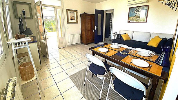 Un coin repas avec une table dressée pour deux personnes, un ventilateur de plafond et une vue sur l’extérieur, visible à travers une porte ouverte.