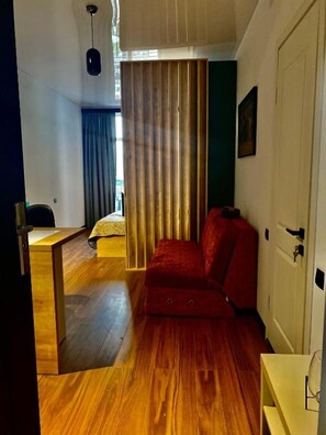 A room with a wooden floor, a red sofa, a bed with a blue curtain, a white door, and a small table with a glass.
