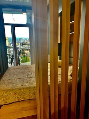 A room with a bed, a wooden partition, and a balcony with a view of buildings and greenery.