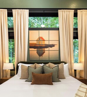 Una habitación de hotel con una cama grande, varias almohadas, un cuadro de una casa en la pared y cortinas.