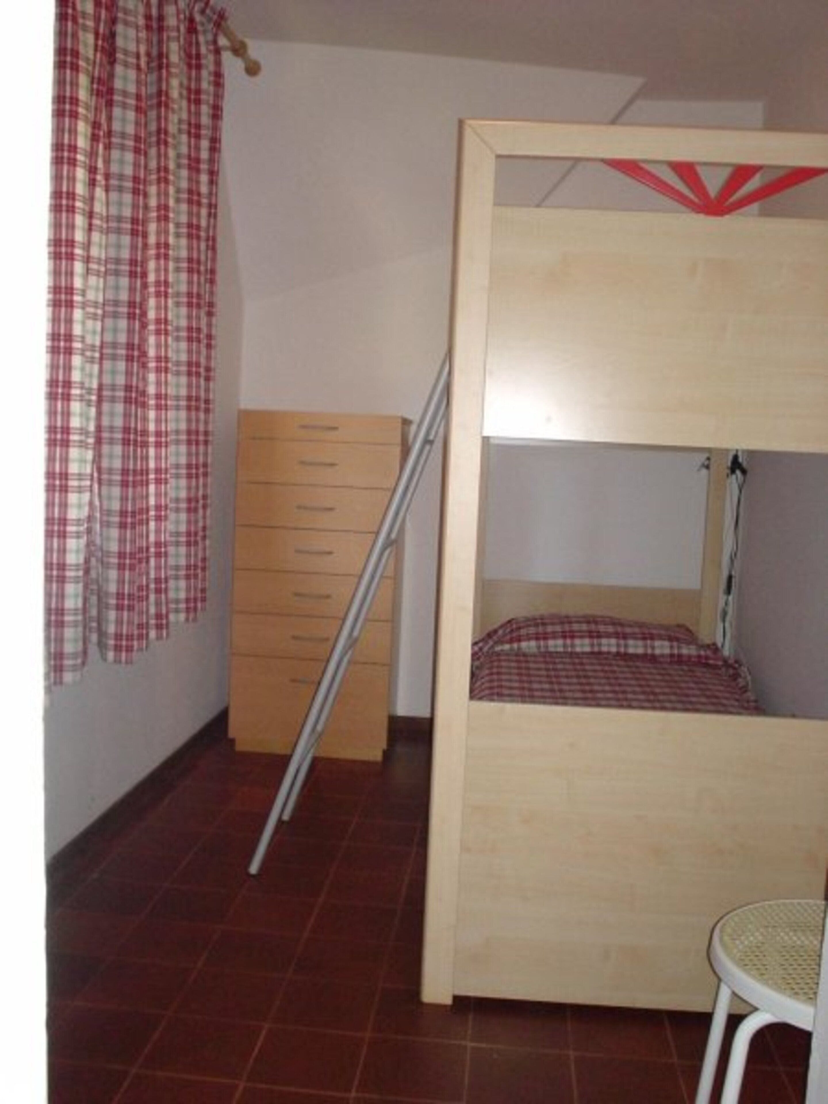 Ein Zimmer mit Etagenbett, einem Holzkasten, einem Stuhl und einem Vorhang.