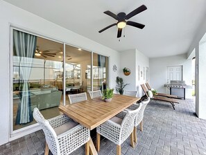 Eine Terrasse mit Holztisch, weißen Korbstühlen, einem Deckenventilator und einem Grillbereich.